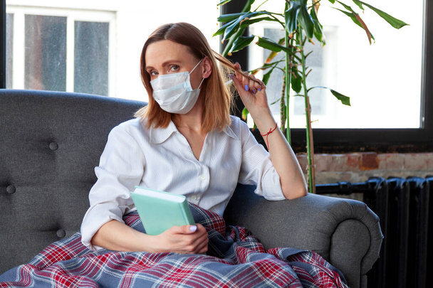 Woman in medical mask holding a book while sitting on a sofa - Photo, Image