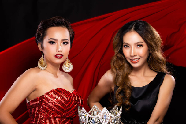 Két Miss Szépségkirálynő versenyző boldog és mosolyog egy gyémánt koronáért. A Sequin Long Ruhát viselik, és díjat adnak a Head-nek a vörös ruha hátterében, ami a levegőben repül. - Fotó, kép