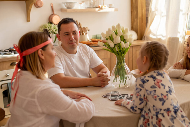 смішні батьки та їхні дочки грають цікаву гру, сидячи на кухні за столом
 - Фото, зображення