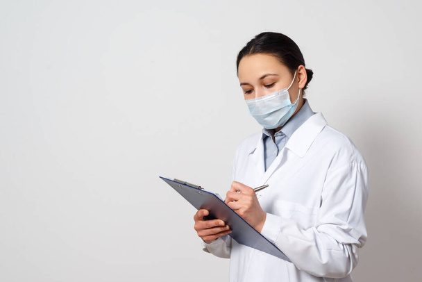 portrait of a female doctor in a protective mask with a clipboard and a pen in her hands, making a record. on white background. - Foto, Imagem
