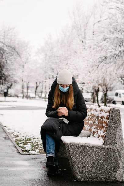 Ένα κορίτσι με αποστειρωμένη ιατρική μάσκα από ιούς και γρίπη κάθεται σε ένα παγκάκι στο πάρκο. Χειμερινή μέρα. Κρύο. Χιόνισμα. - Φωτογραφία, εικόνα