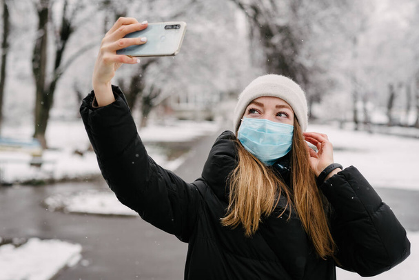 Egy lány egy steril orvosi maszkban vírusokból, és az influenza leveszi magát egy mobiltelefon kamerával. Havazik odakint. Emberek nélkül. Homályos háttér - Fotó, kép