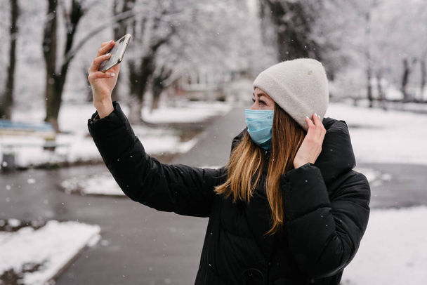 Een meisje met een steriel medisch masker van virussen en de griep neemt zichzelf af met een mobiele telefoon camera. Het sneeuwt buiten. Zonder mensen. Wazige achtergrond - Foto, afbeelding