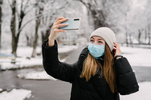 Een meisje met een steriel medisch masker van virussen en de griep neemt zichzelf af met een mobiele telefoon camera. Het sneeuwt buiten. Zonder mensen. Wazige achtergrond - Foto, afbeelding