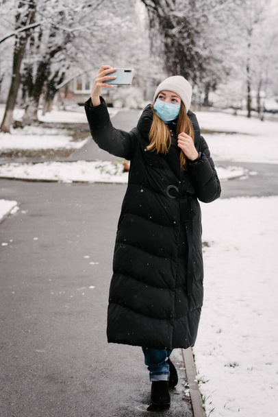 Dziewczyna w sterylnej masce medycznej od wirusów i grypa zdejmuje się z aparatu telefonicznego. Na zewnątrz pada śnieg. Bez ludzi. Zamazane tło - Zdjęcie, obraz