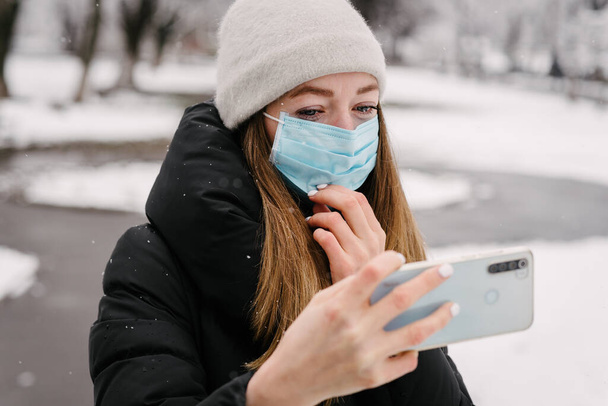 Une fille dans un masque médical stérile contre les virus et la grippe s'enlève sur un appareil photo de téléphone portable. Il neige dehors. Sans personne. Fond flou
 - Photo, image