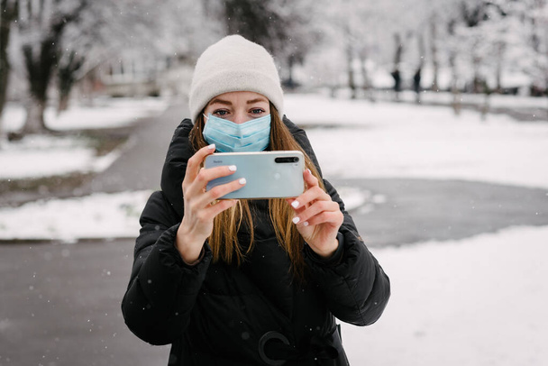 Steril tıbbi maske takmış virüslü bir kız ve grip cep telefonu kamerasından kendini çıkarıyor. Dışarıda kar yağıyor. İnsanlar olmadan. Bulanık arkaplan - Fotoğraf, Görsel