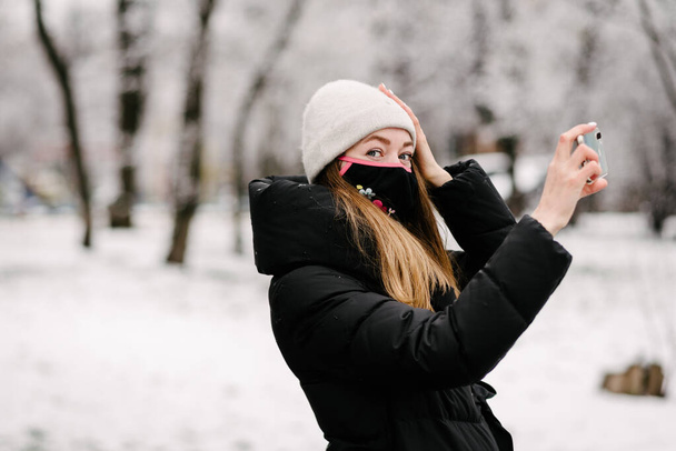 Una chica con una máscara médica estéril de virus y la gripe se quita en una cámara de teléfono móvil. Está nevando afuera. Sin gente. Fondo borroso
 - Foto, Imagen
