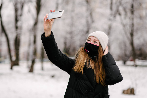 Uma garota com uma máscara médica estéril de vírus e a gripe se descola em uma câmera de telefone celular. Está nevando lá fora. Sem pessoas. Fundo desfocado
 - Foto, Imagem