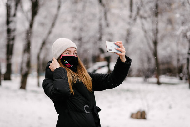Dziewczyna w sterylnej masce medycznej od wirusów i grypa zdejmuje się z aparatu telefonicznego. Na zewnątrz pada śnieg. Bez ludzi. Zamazane tło - Zdjęcie, obraz
