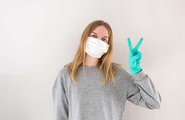 Een blonde vrouw die onderzoek doet met een medisch masker maakt vrede of overwinningsteken. Het concept van de epidemie van het coronavirus en anti-smog. Corona virus 2019-ncov covid-19. Mensen en lichaamstaal. - Foto, afbeelding