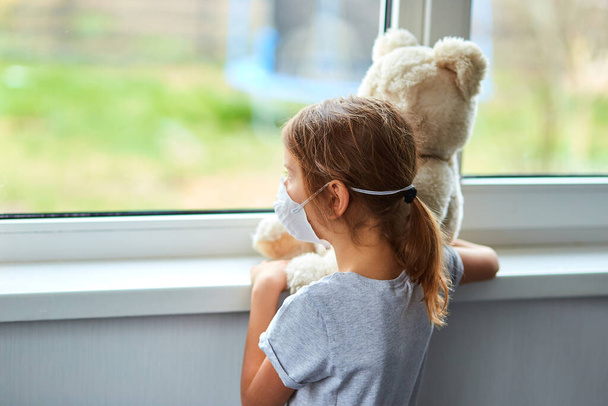Little girl holding and hugging teddy bear in mask near the window. Coronavirus, covid-19 and pollution protection concept. Stay at home. - Foto, Imagem