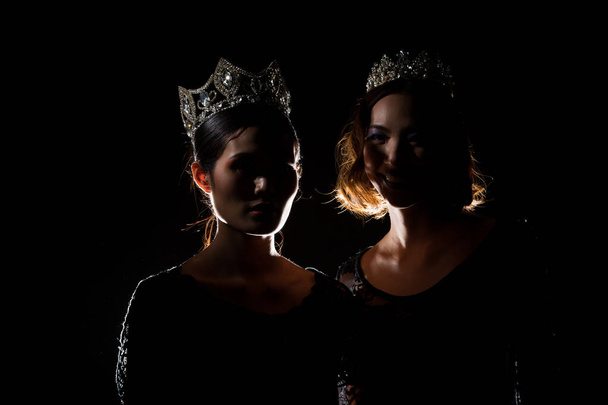 Two Silhouette Shadow Back Rim Light of Miss Pageant Beauty Queen Contest with Silver Diamond Crown stand together, studio lighting dark black background, multinational asian and caucasian models - Photo, Image