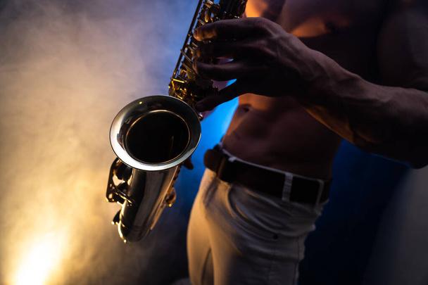 Muscular man with naked torso playing on saxophone with smoked colorful background  - Foto, imagen