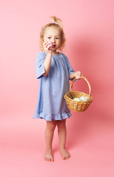 Feliz Pascua: una niña con un vestido azul sostiene una canasta de huevos en sus manos. Retrato sobre fondo rosa
. - Foto, imagen