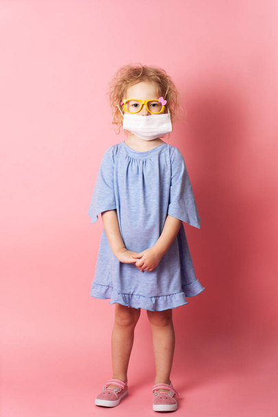 Children's medicine: a little girl in a blue dress and in a medical mask holds. Portrait on a pink background. - Foto, immagini