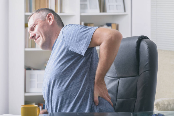 Hombre en la oficina del hogar que sufre de dolor de espalda baja sentado en un escritorio con cuaderno, papeles y otros objetos
 - Foto, imagen
