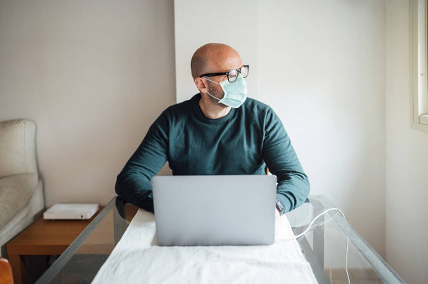 Hombre trabajando desde casa en un ordenador portátil sentado en un escritorio con máscara
 - Foto, Imagen