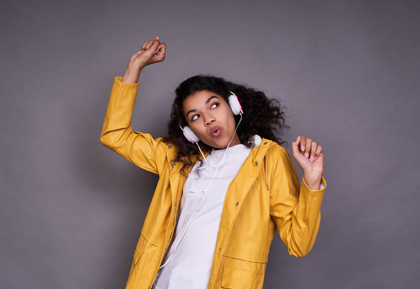 A cheerful young dark-skinned girl, in a white T-shirt and a yellow jacket, with magnificent curly hair, listens to music on headphones, sings, dances with her favorite music, looks to the side. - Zdjęcie, obraz