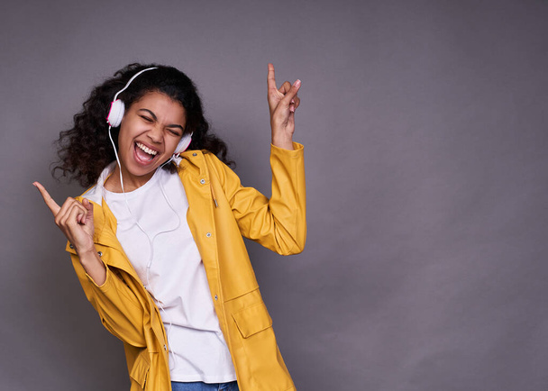 An expressive young dark-skinned woman in white T-shirt and yellow jacket, with magnificent curly hair, listens to music on headphones, shows the peace sign with her hands, enjoys her favorite music. - Zdjęcie, obraz