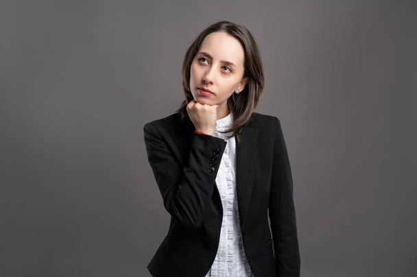 Portrait of wonderful young business woman wering black suit and shirt holding hand under her chin, having doubtful poseing on isolated gray background - Foto, Imagen