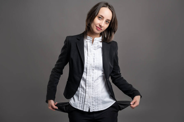 Porträt einer wunderbaren jungen Geschäftsfrau in schwarzem Anzug und Hemd, die leere Taschen vor isoliertem grauen Hintergrund posiert - Foto, Bild