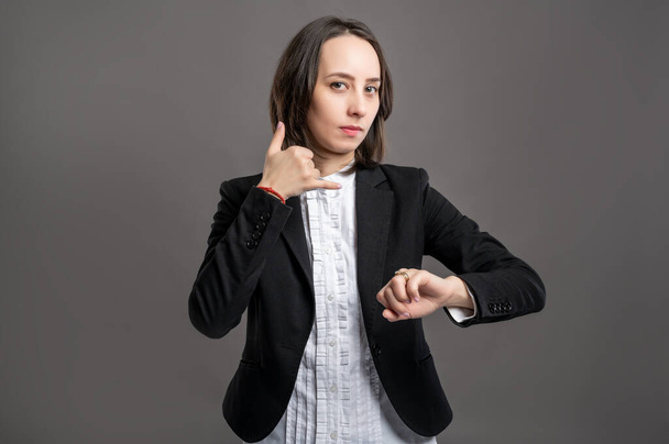 Portrait of wonderful young business woman wering black suit and shirt showing call me gesture and looking her watch poseing on isolated gray background - Foto, imagen