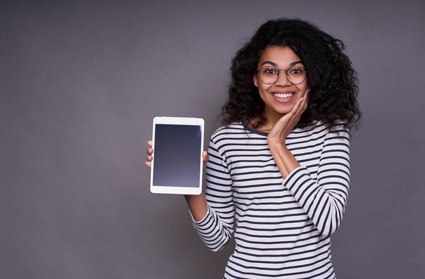 A pretty surprised sweet young African American girl in a striped sweater and glasses, with curly black hair, holds a tablet in her right hand and touches her face with her left hand. - Foto, imagen