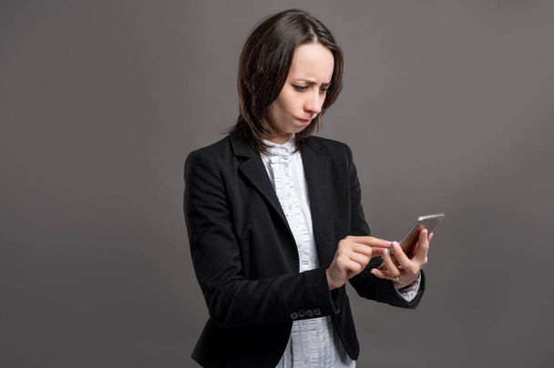 Портрет замечательной молодой деловой женщины в черном костюме и рубашке, держащей смартфон, смс, выглядящей злой, позирующей на изолированном сером фоне
 - Фото, изображение