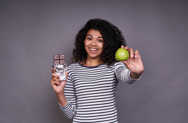 Cute African woman in a striped sweater, with a lush curly hairstyle, holds a green apple and a chocolate bar in her hand, looks straight with smile and shows apple on frame. - Photo, Image