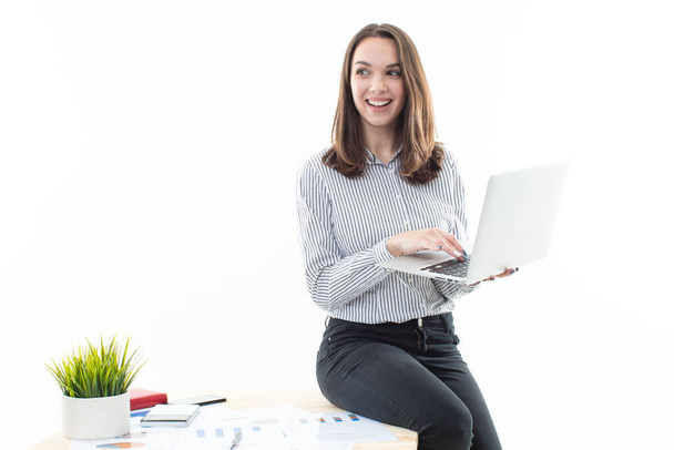 A girl in a good mood is sitting on a table and typing on a computer on a white background - Photo, Image