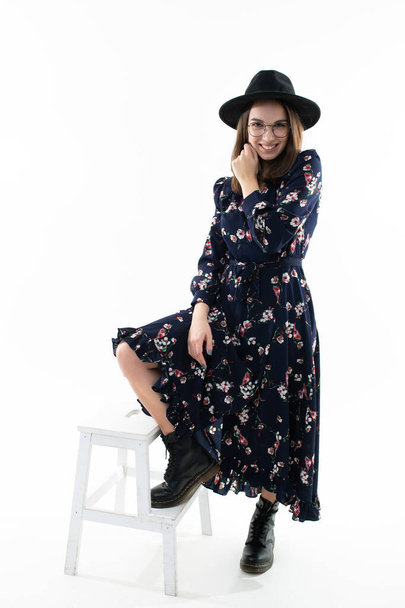 A brunette in a long floral dress with a hat poses for the camera in the studio. Emotional portrait - Foto, Imagem