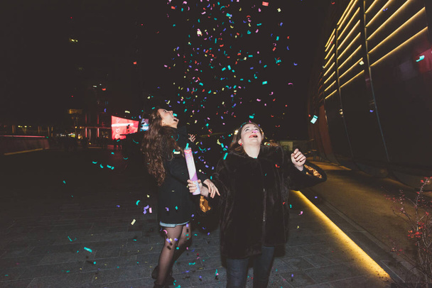 Deux jeunes belles femmes caucasiennes amies en plein air dans la nuit de la ville jouant avec des confettis colorés - célébration, événement, concept de fête
 - Photo, image