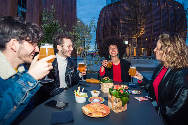 Group of four multiethnic friends sitting outdoor club drinking beer making toast - social gathering, togetherness, celebration concept - Photo, Image