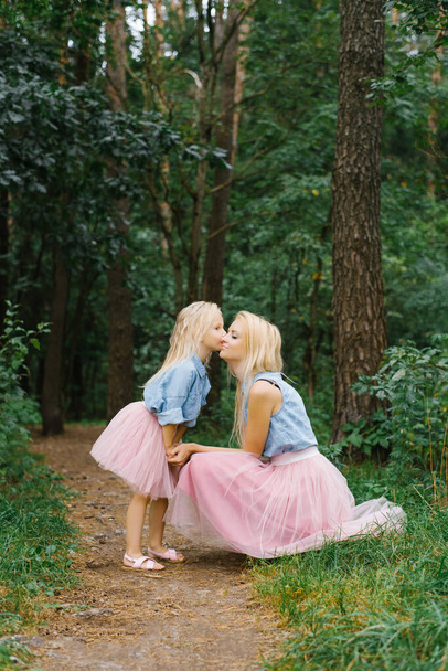 Egy anya és egy ötéves lány ugyanabban a romantikus ruhában sétálnak a parkban vagy az erdőben. A lányom arcon csókolja anyámat. - Fotó, kép