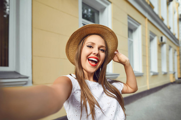 A girl with a beautiful smile in a hat makes a selfie on the phone on a city street. - Zdjęcie, obraz