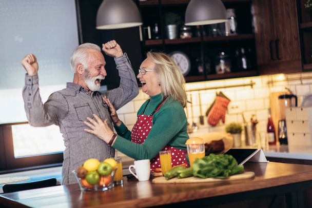 Beautiful senior couple is dancing and smiling while cooking together in kitchen. Quarantine. Health concept. - 写真・画像
