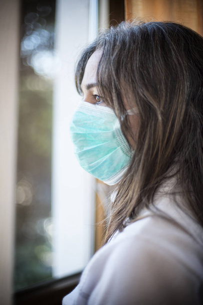 Wirus korony. Chora kobieta z wirusem korony patrząca przez okno i nosząca bezpieczną ochronę przed oddychaniem i odzyskiwaniem sił po chorobie w domu. Izolowany pacjent w celu zapobiegania zakażeniom - Zdjęcie, obraz