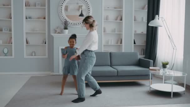 Caring mother having fun and cool time dressed in white shirt dress concept multinational family gorgeous joyful mother jumping funny kid african daughter dancing together in apartment with sofa - Footage, Video