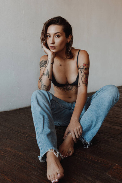 Sexy woman with tattoos wearing lingerie and jeans sitting on the floor - Photo, image