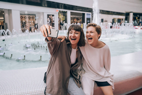 İki kız alışveriş merkezinde selfie çekiyor, arka planda bir çeşme. - Fotoğraf, Görsel