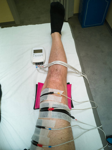 Нога человека с устройством электростимуляции мышц и ледяной мешок во время восстановительных упражнений на кровати после операции на колене
. - Фото, изображение