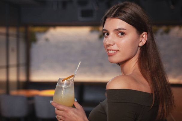 Κοντινό πλάνο μιας όμορφης νεαρής γυναίκας που χαμογελάει πάνω από τον ώμο της στην κάμερα, απολαμβάνοντας ένα ποτό στο μπαρ - Φωτογραφία, εικόνα