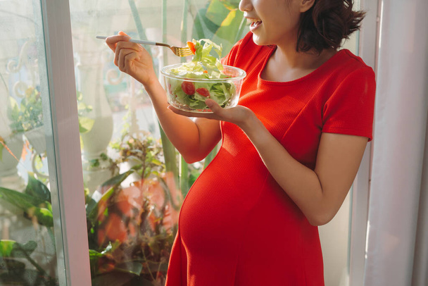 Jovem grávida comendo salada de legumes perto da janela em casa
 - Foto, Imagem