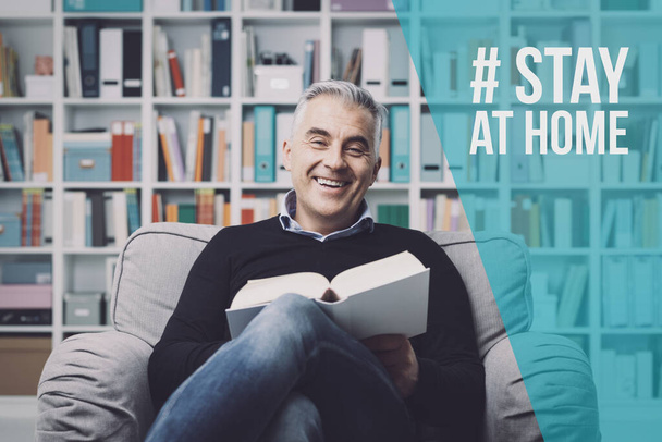Smiling man reading a book and relaxing at home alone: stay at home social media campaign for coronavirus prevention - Photo, image