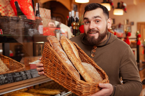 Genç sakallı adam kameraya gülümsüyor, elinde bir sepet dolusu ekmek var. Erkek müşteri yerel fırından alışveriş yapıyor. - Fotoğraf, Görsel