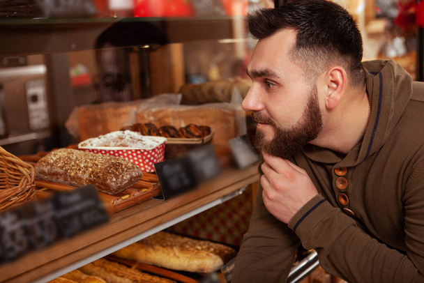Großaufnahme eines bärtigen jungen Mannes, der sich ein Dessert aussucht, das er an der Auslage in der Bäckerei kaufen kann - Foto, Bild