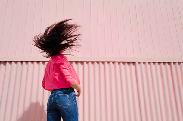 Young woman stands her back to camera and waving her long straight dark hair on pink background. Female with flying hair in pink leather jacket and blue jeans against colored iron wall. Copy space - Photo, image