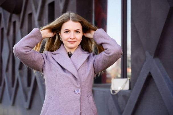 Portrait of a blonde Caucasian girl with long beautiful hair outdoors in a pink coat against the background of a building window in the afternoon on a sunny day. - 写真・画像