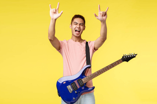 Hezký mladý hudebník hrající na kytaru a zpěv, izolovaný na žlutém pozadí - Fotografie, Obrázek